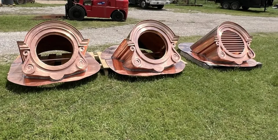 Three copper chimney caps with louvered sides lined up on a lawn before installation