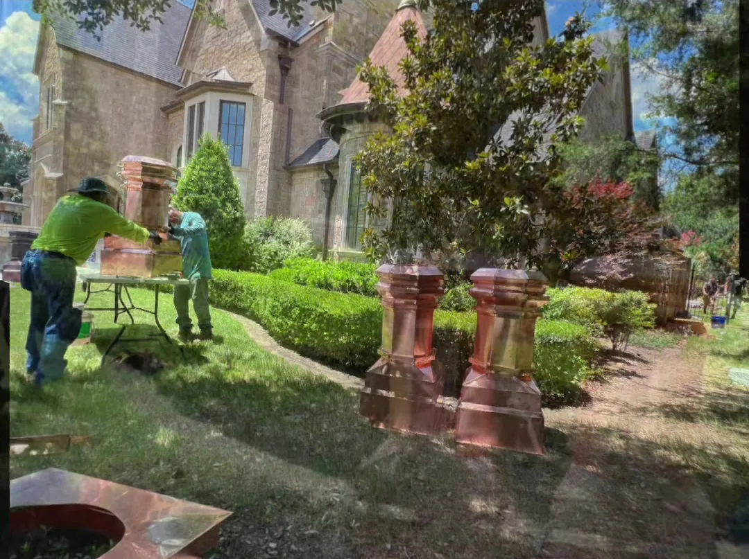 Two crew members at a folding table on a lawn beside a stone house, with two finished copper chimney caps standing on the grass