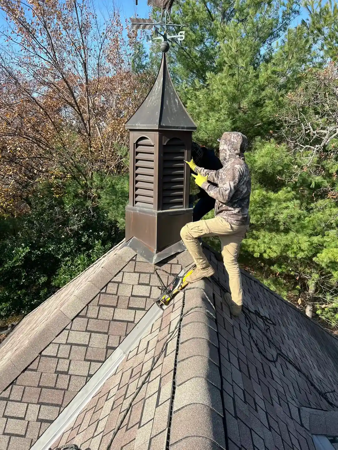 A crew member on a shingled roof fitting a louvered copper cupola, trees behind