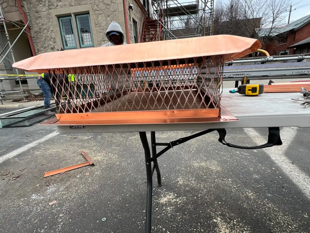 Copper chimney cap with a hipped lid and a diamond-pattern screen, on a folding table outside