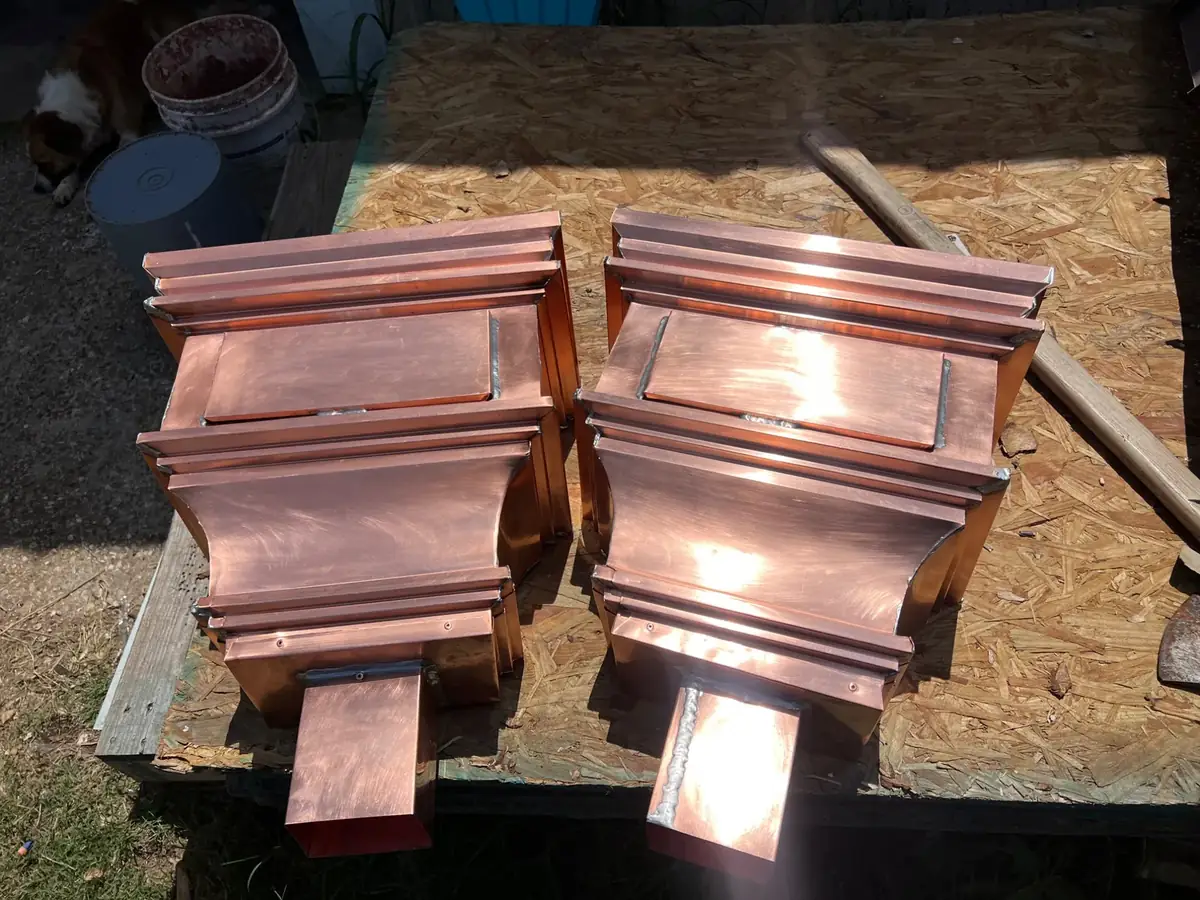 Two copper collector heads with stepped crown bands and rectangular outlets, on a plywood bench