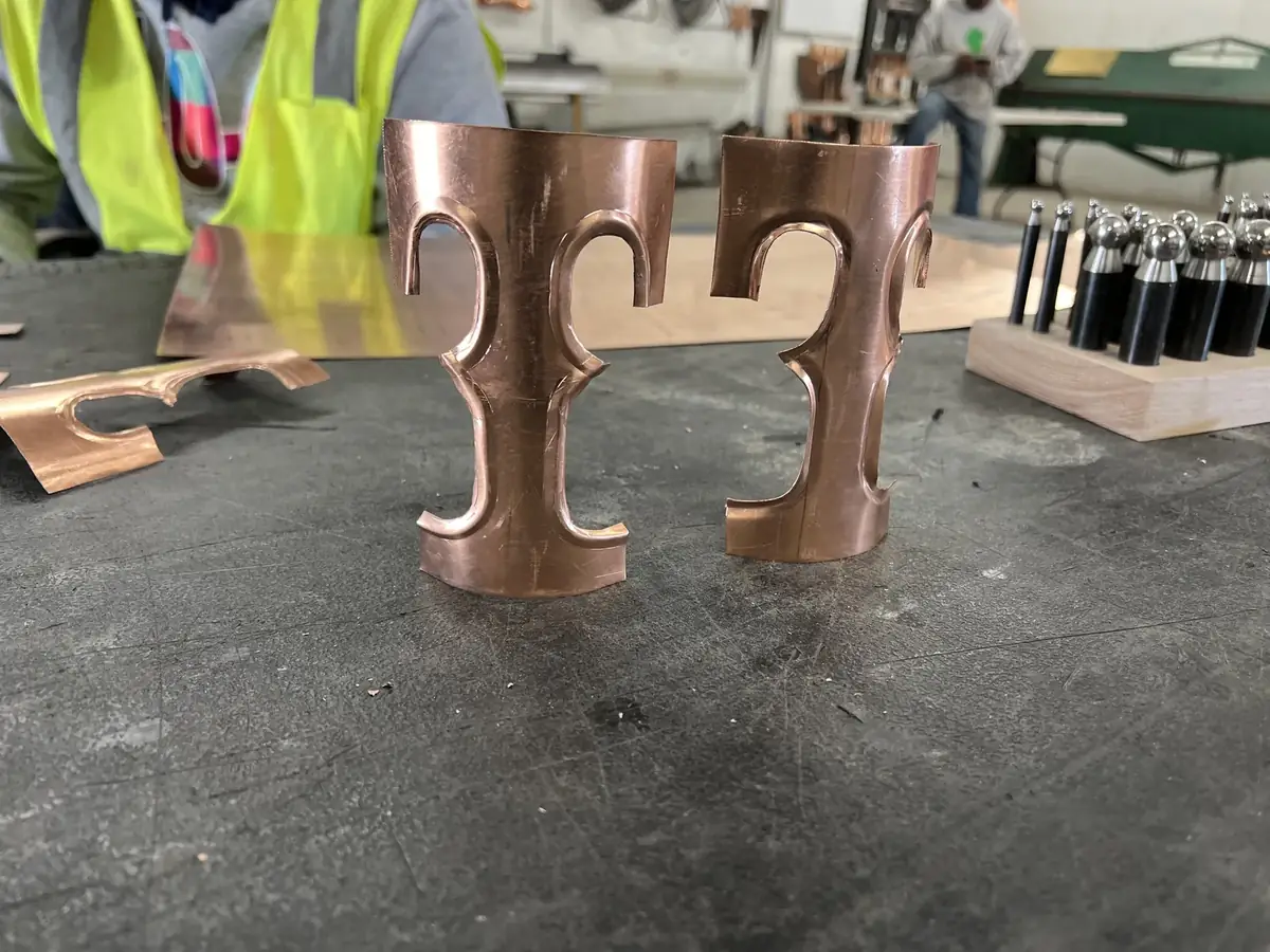 Two formed copper downspout bands with curled, scrolled arms, standing on a shop bench