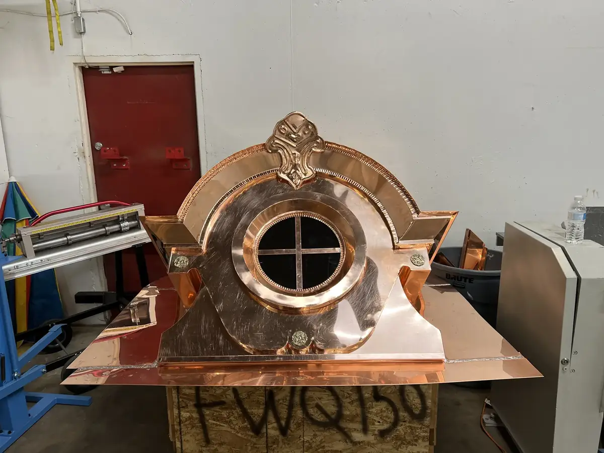 Copper oval-window dormer with a round sash, scrolled trim and a crest ornament, on a crate in the shop