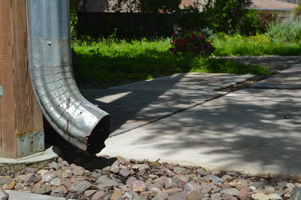 The foot of a galvanized steel downspout turning out over a bed of river stones beside a path