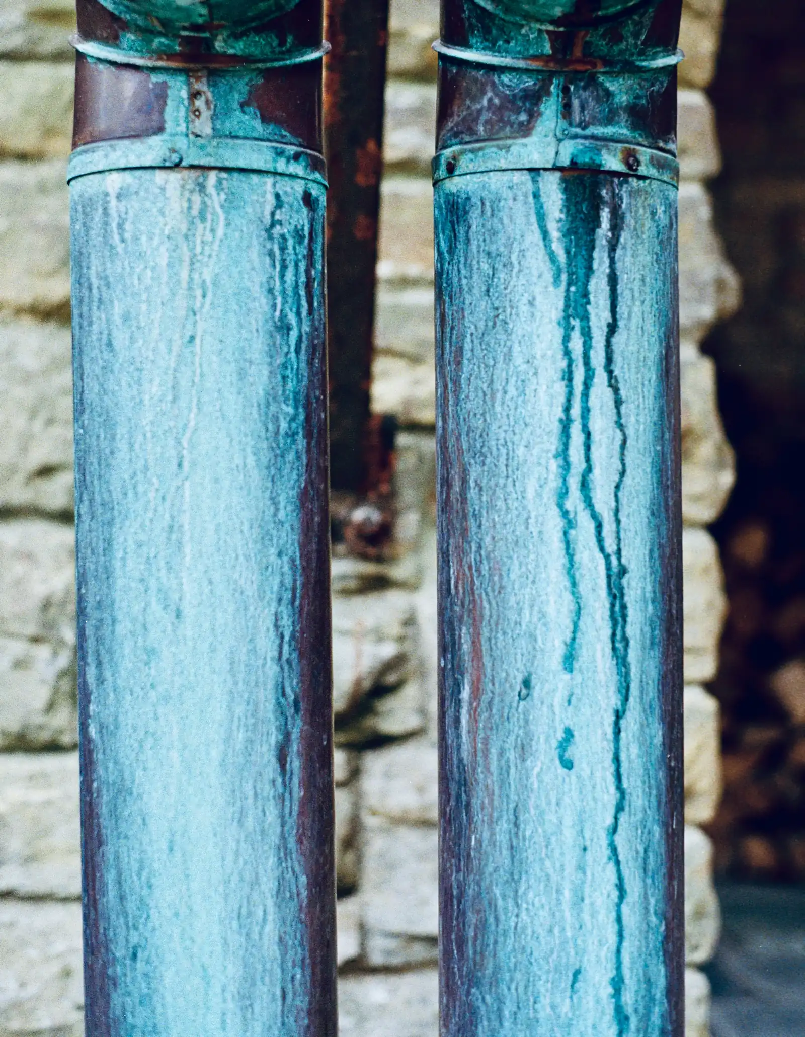 Two copper downspouts weathered to a blue-green patina, against a stone wall