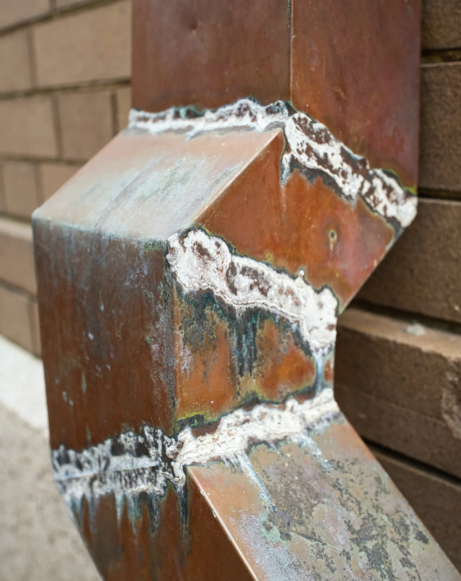 Rectangular copper downspout with soldered offset joints, weathered to brown with streaks of green